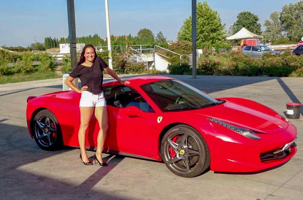 A woman dressed nice in short white shorts, black shirt and high heels next to a red Ferrari to fulfill her Italy bucket list.