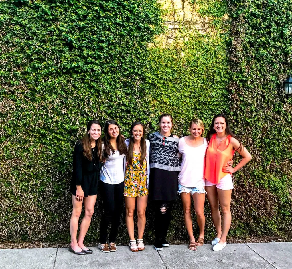 Six girls enjoying their girls weekend in Fredericksburg, Texas, smiling against a wall covered in vines. 