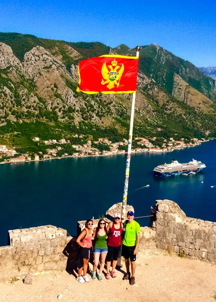 A group of four (three girls and one guy) standing at Kotor's fortress with the Montenegro flag towering over them and the Bay of Kotor with a cruise ship in the background. 