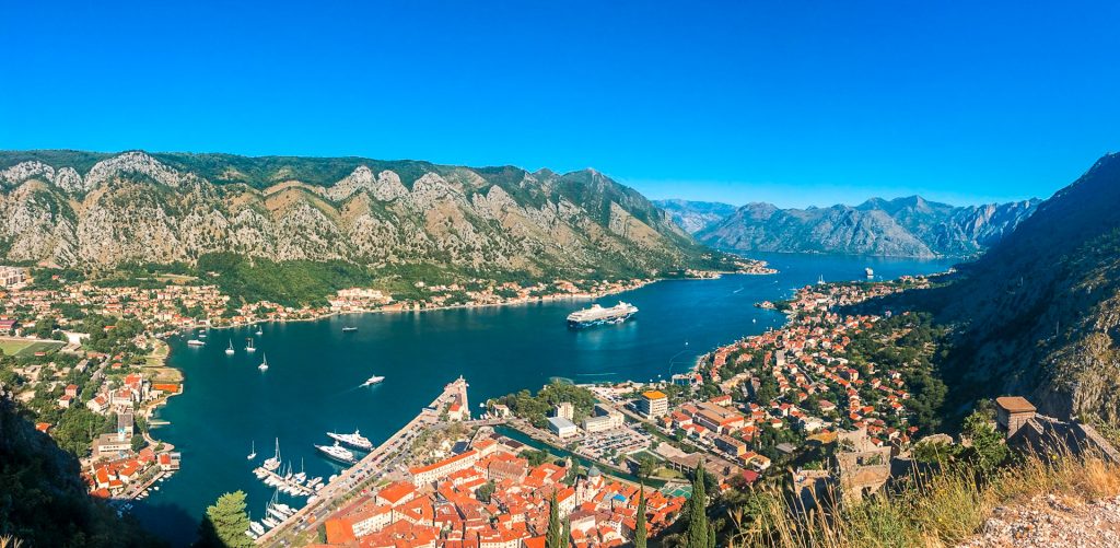 Panorama picture from the top of Kotor's castle - the Castle of San Giovanni looking down at the Bay of Kotor and Old Town Kotor in the shape of a triangle leading into the deep blue ocean with mountains hovering over. 