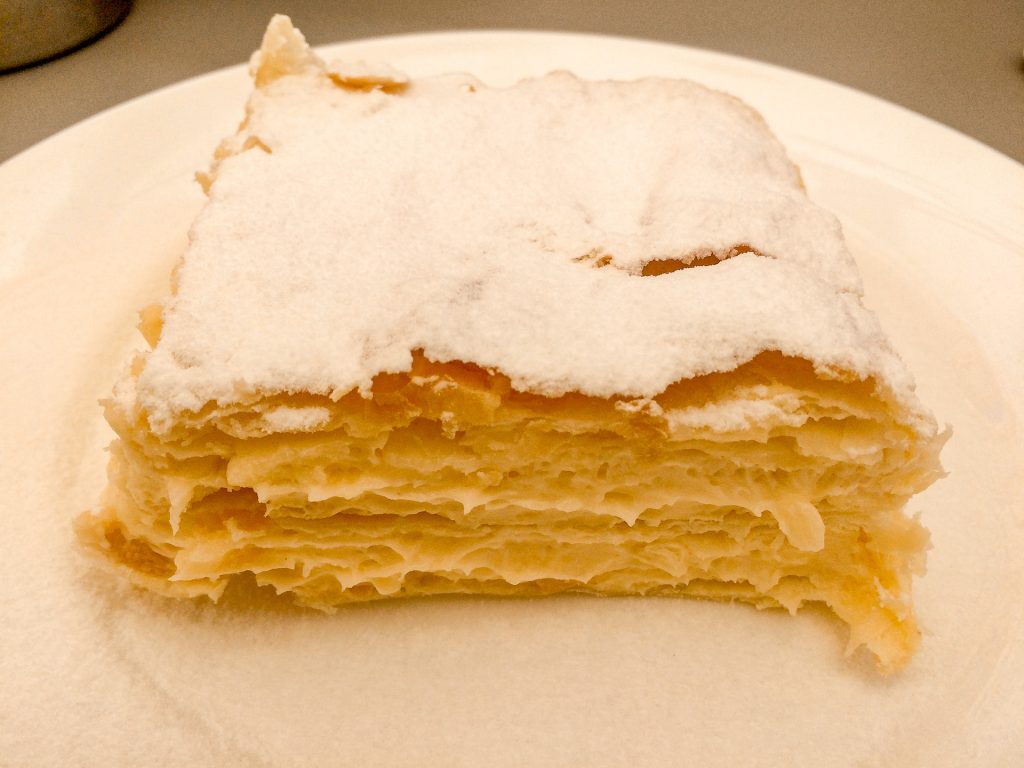A square piece of krempita with sweet cream filled between layers of flaky puff pastry dough and covered in powdered sugar. 
