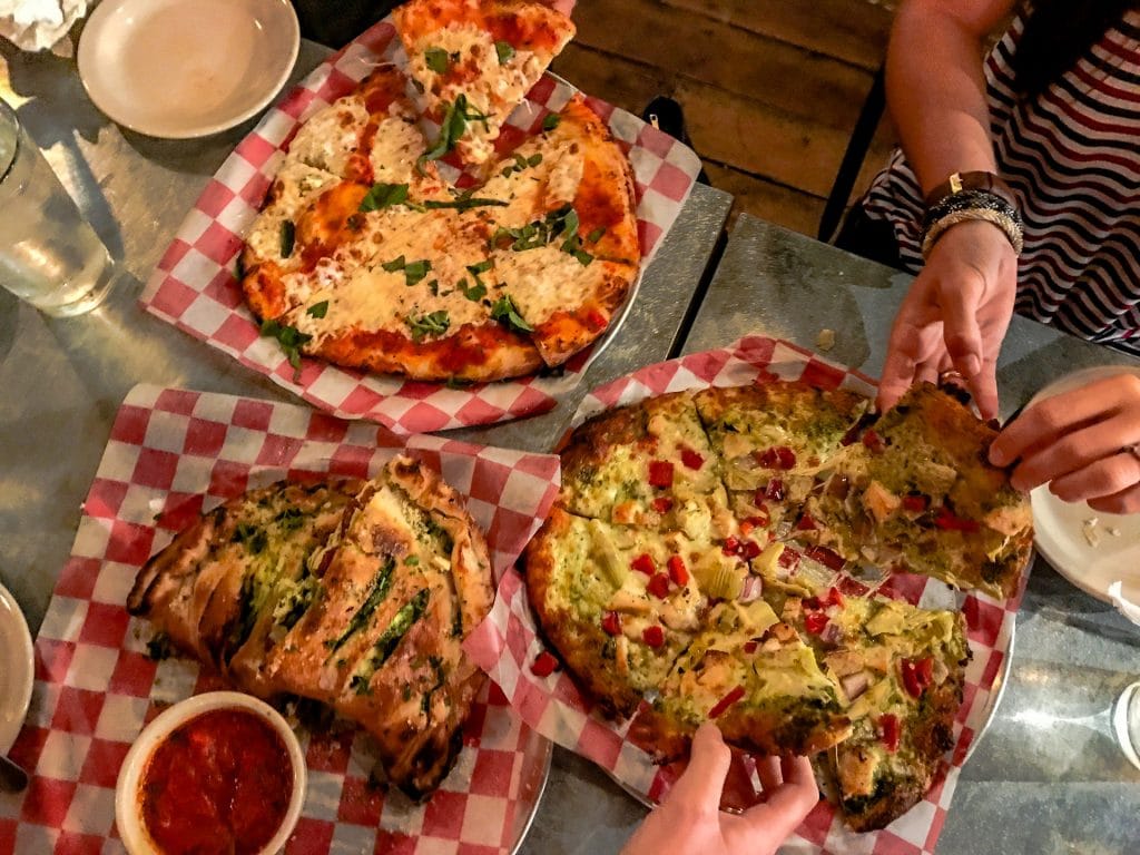 Hands taking a slice from two whole pizzas and a calzone at Woerner Warehouse.
