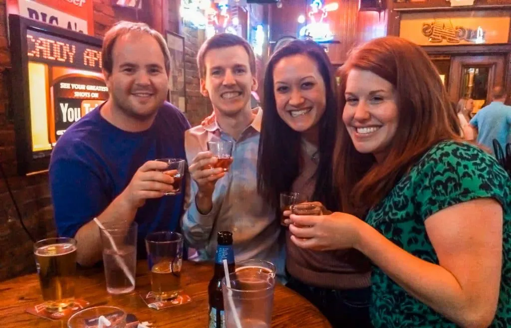 Four friends smiling for a photo and toasting with shot glasses at Big A's.