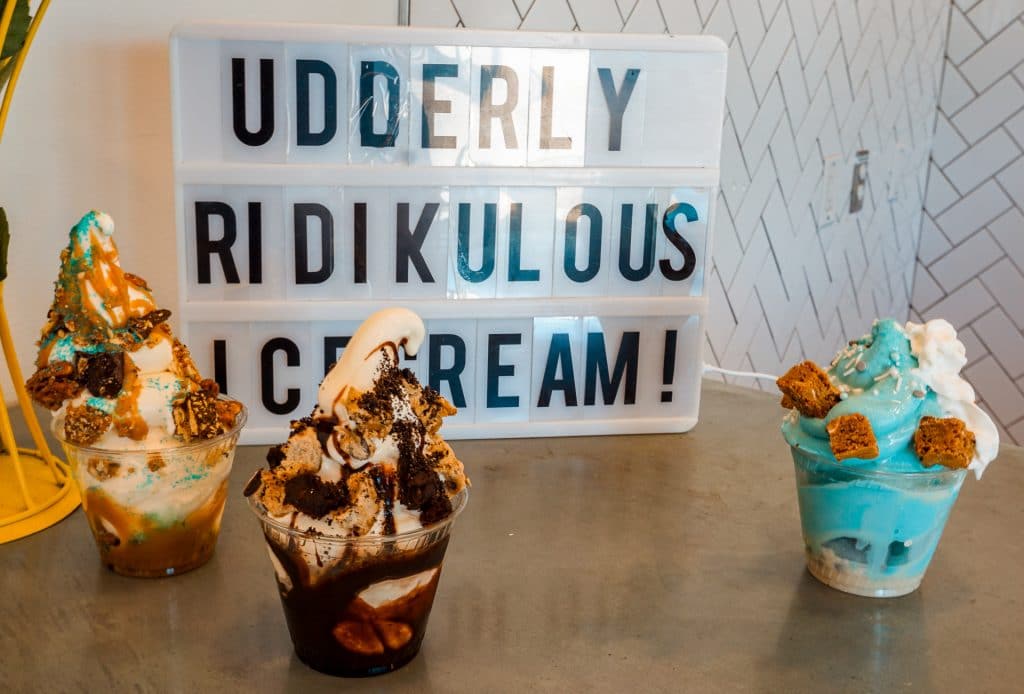 Three different soft serve ice cream sundaes with a sign in the background that says "UDDERLY RIDIKULOUS ICE CREAM!" from Cow Tipping Creamery in Carrollton.