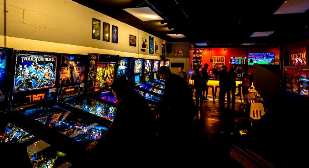 A dark room with people playing classic pinball machines at Two Plumbers Arcade and Brewery.