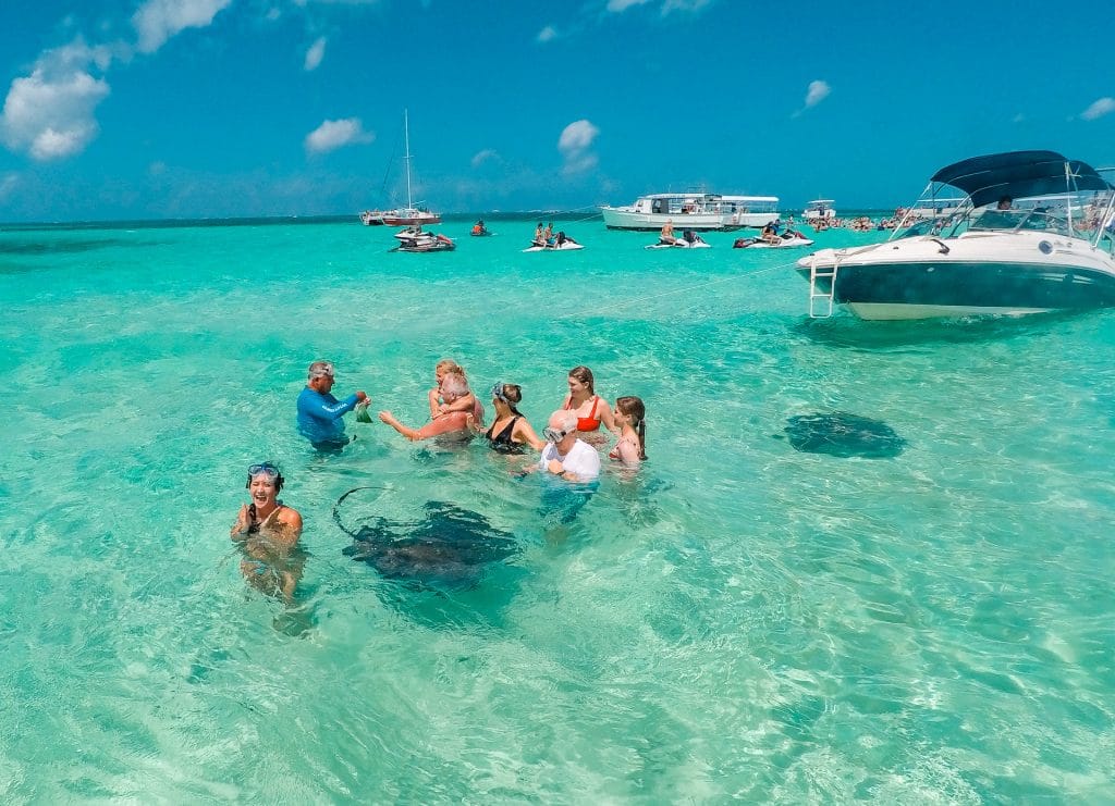a group of people standing in shallow water with stingrays swimming nearby. There are also more people and boats in the background. 