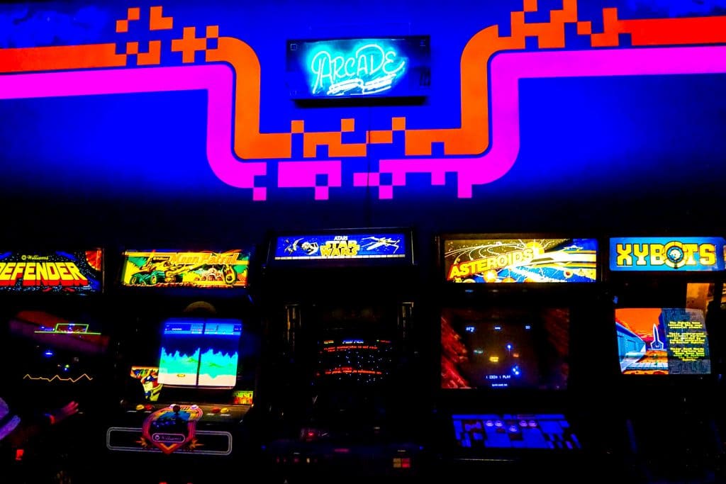 A bright blue and pink neon wall with a row of classic arcade games lined up located at the National Video Game Museum in Frisco.