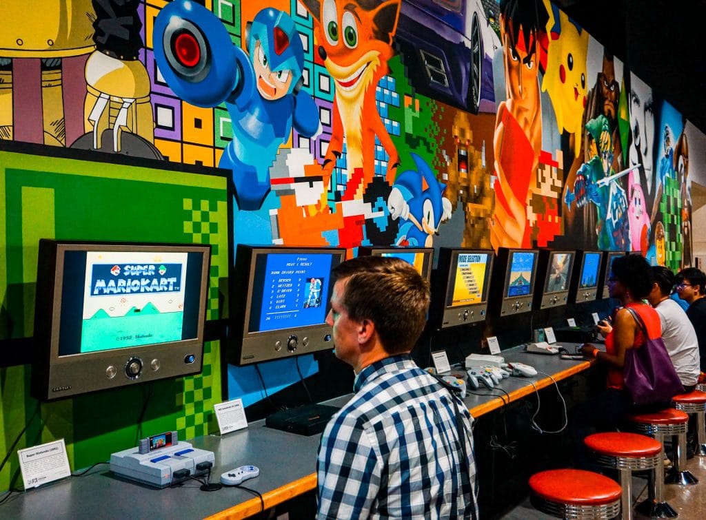 A man playing Nintendo Classic with a row of other gaming consoles and a video game mural on the wall at the National Video Game Museum in Frisco. 
