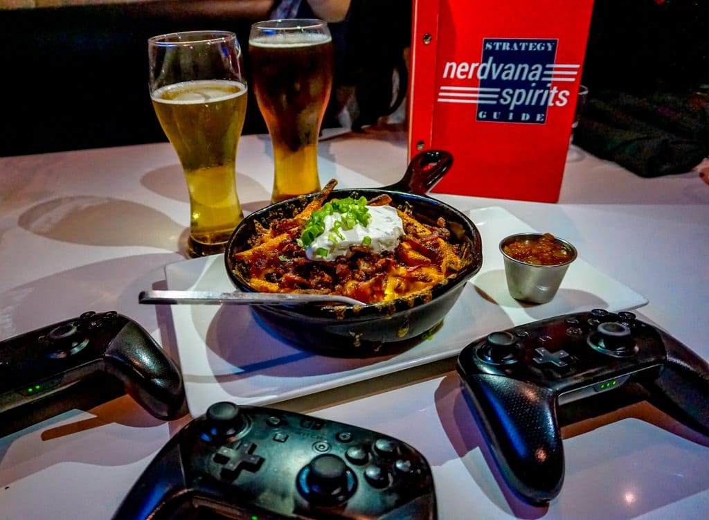 A table with three black Switch controllers,  a skillet of loaded fries and two glasses of beer from Nerdvana.