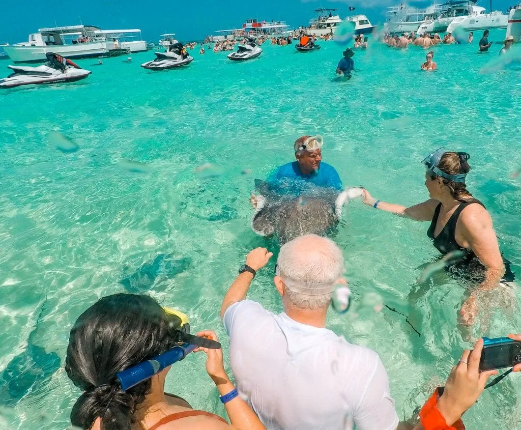 A few people gathered around stroking a stingray at Stingray City with more people and boats in the distance. 