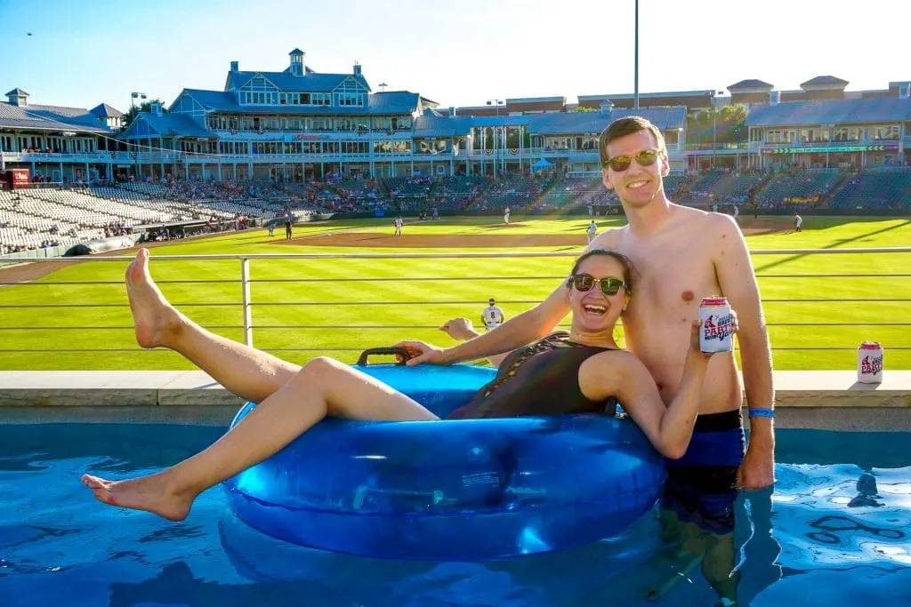 A woman laying inside a blue float with a beer in her hand and a man standing next to her in a lazy river overlooking the RoughRiders baseball game. 