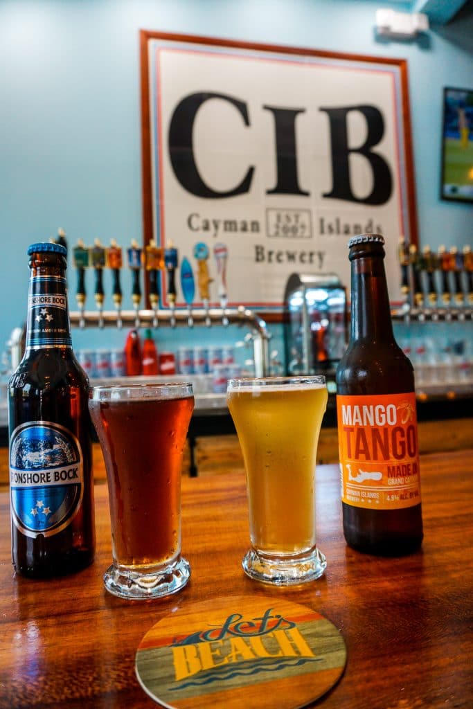 two glasses and bottles of beer on a wooden bar top with Cayman Islands Brewery logo in the background.  