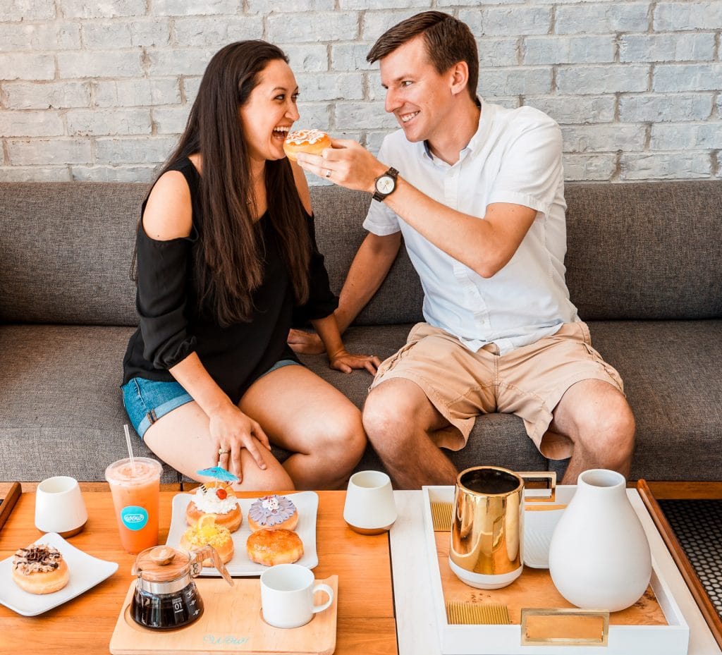A man feeding a woman an orange glazed donut. The table in front of them has coffee, tea, and a plate full of donuts.