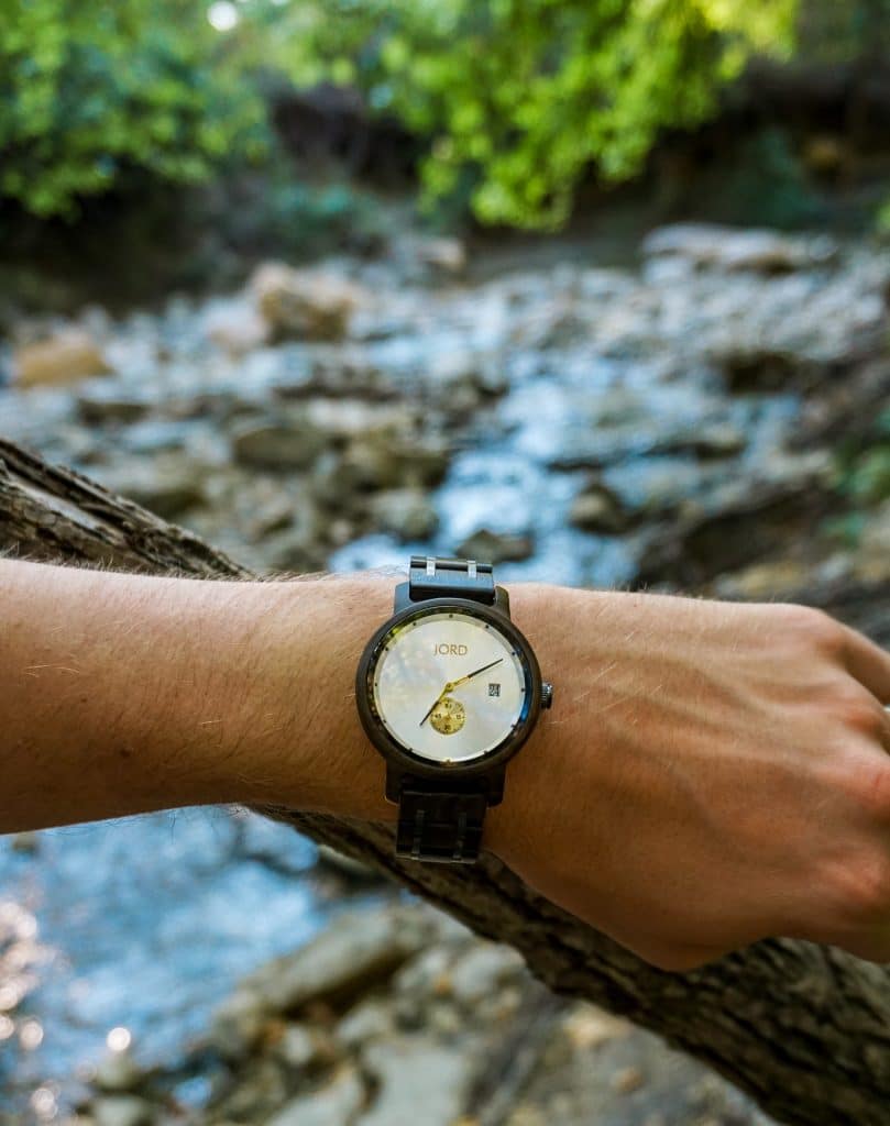 A man's arm in the center of the photo representing a wooden watch from JORD with a stream of water in the background.