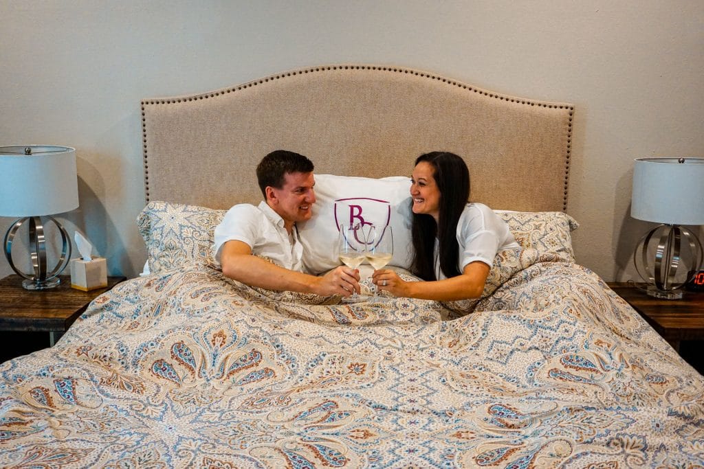 A couple toasting wine glasses in a comfy bed.