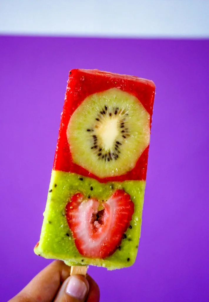 A frozen paleta. The bottom half is kiwi with a sliced strawberry and the top half is strawberry with a sliced kiwi.