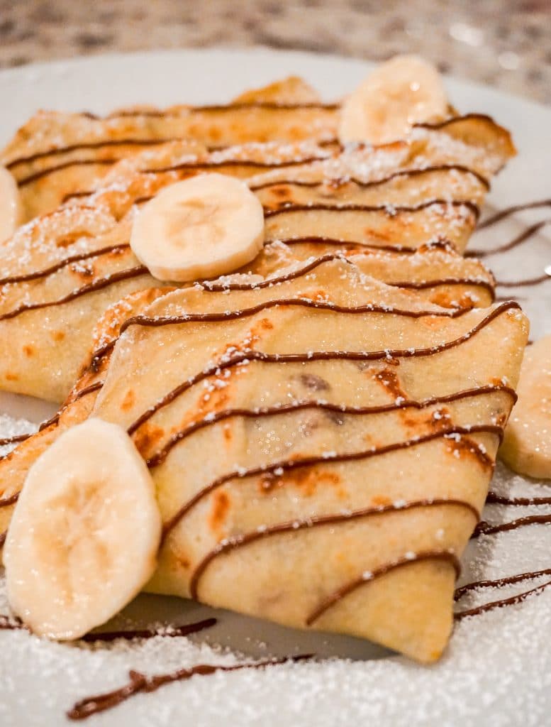 A close up photo of triangular sweet crepes with banana and Nutella filled inside. Then on top of the crepes are fresh sliced bananas, powdered sugar, and drizzled Nutella.