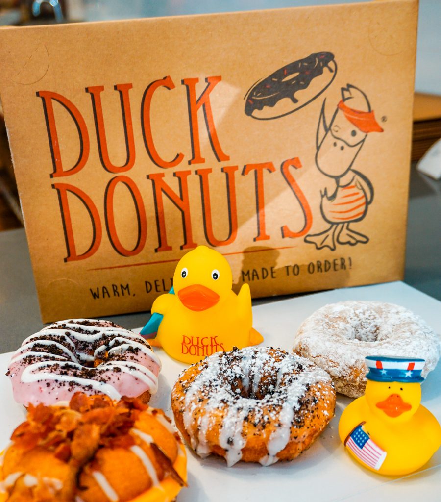 Powdered sugar and glazed cake donuts with little yellow rubber ducks next to the donuts. A cardboard box background with Duck Donuts on it.