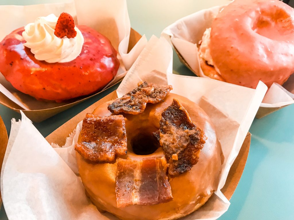 A close up of a maple glazed donut with bacon on top. Two pink donuts are in the background.