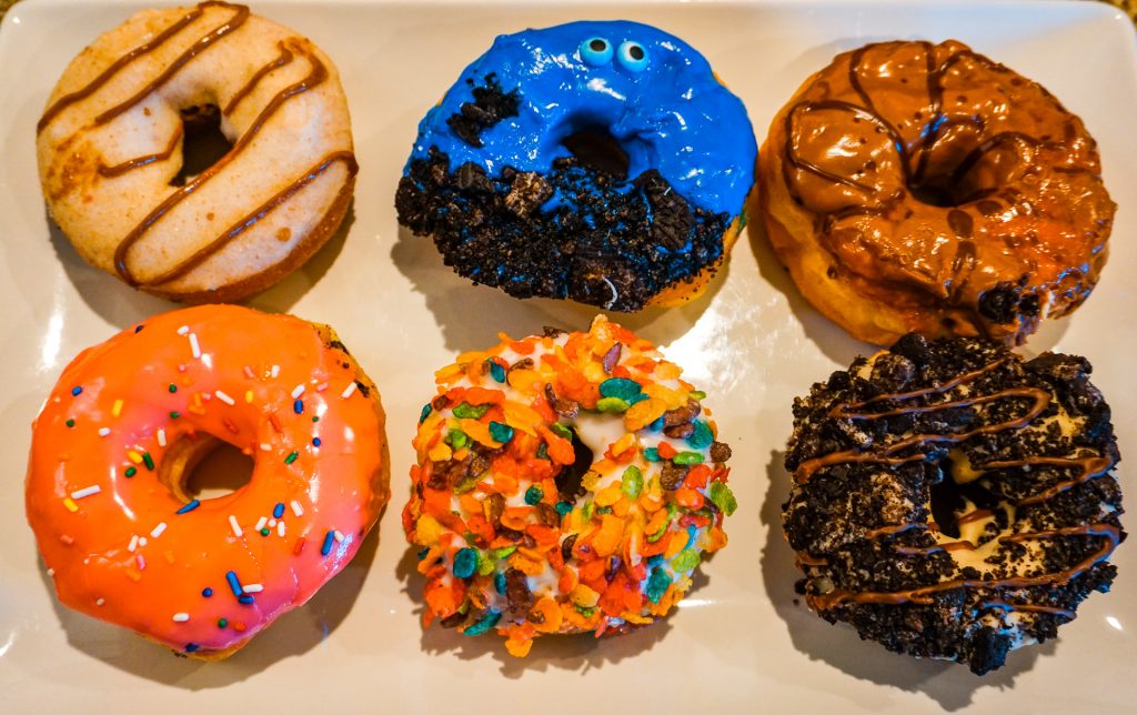 A variety of six of the best donuts in Dallas from Hurts Donuts. There are donuts with cinnamon sugar, a blue donut with chocolate cookie crumbs, a caramel icing, pink icing with sprinkles, Fruity Pebbles, and chocolate cookies covered on top. 
