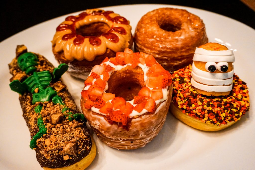 A plate full of some of the best donuts in Dallas. These scrumptious looking donuts include croissant donuts covered in peanut butter and jelly, and strawberries. Then there are Halloween decorated donuts that have a Twinkie mummy and a green skeleton. These donuts can be found at Jarams Donuts.