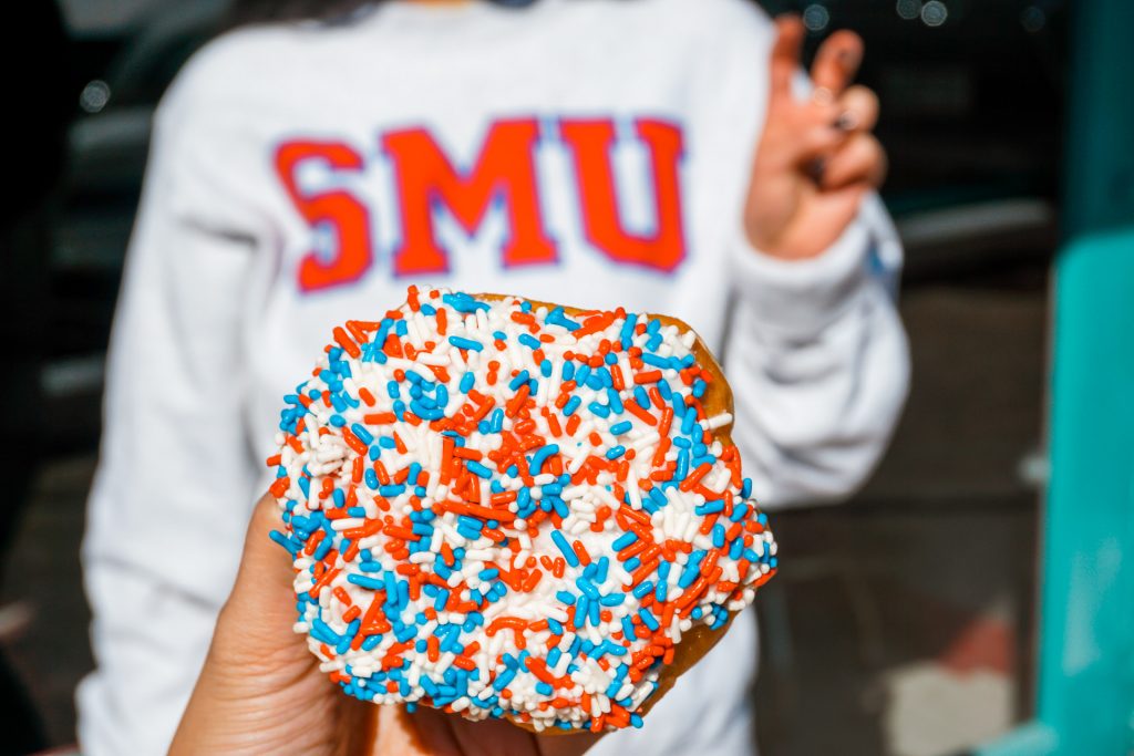 A close up of a red, white, and blue sprinkled donut with a girl in the background with a SMU sweatshirt.