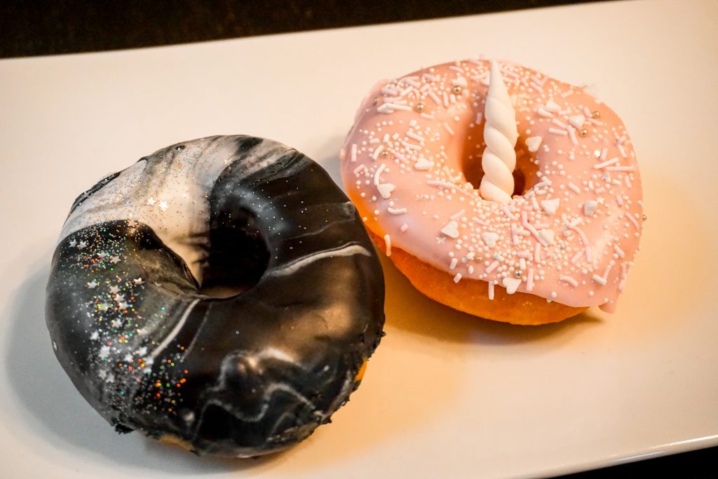 A black and white galaxy with sparkly sprinkles and a pink donut with unicorn horn on top of yeast donuts from Sweet Daze Dessert Bar.