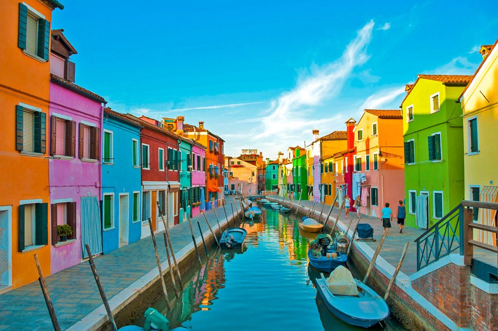 A small canal filled with boats in-between two rows of the most colorful houses in Burano, Italy.