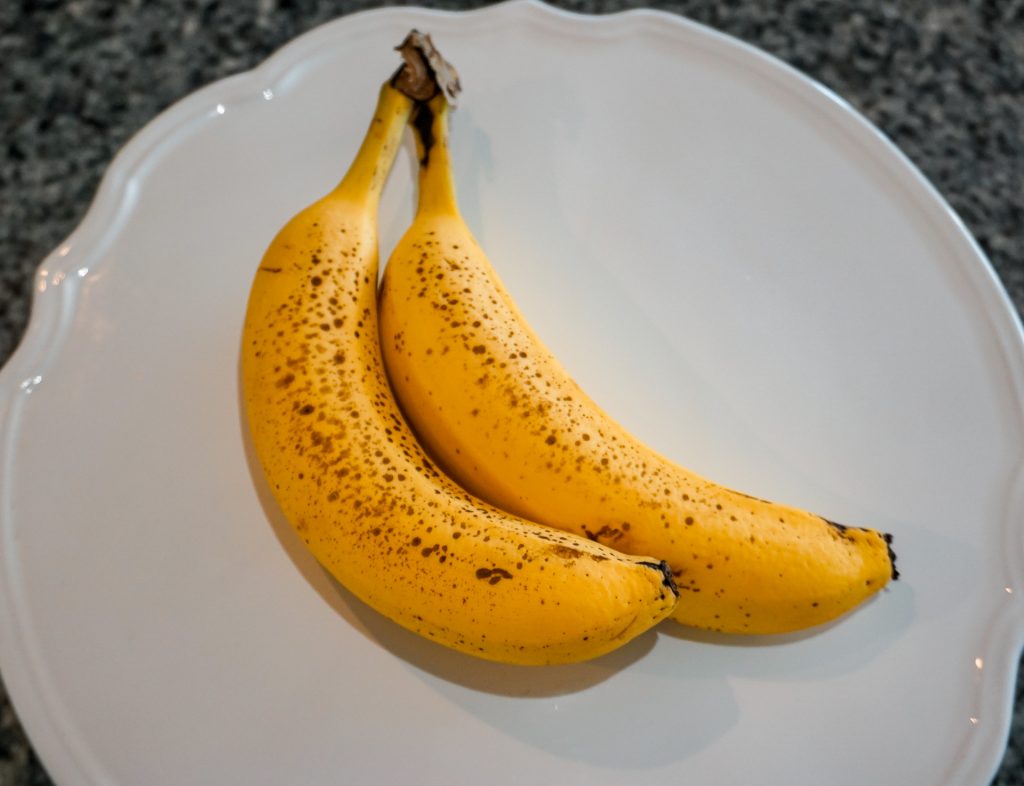 Two ripe bananas - bright yellow with brown spots, on a white plate.