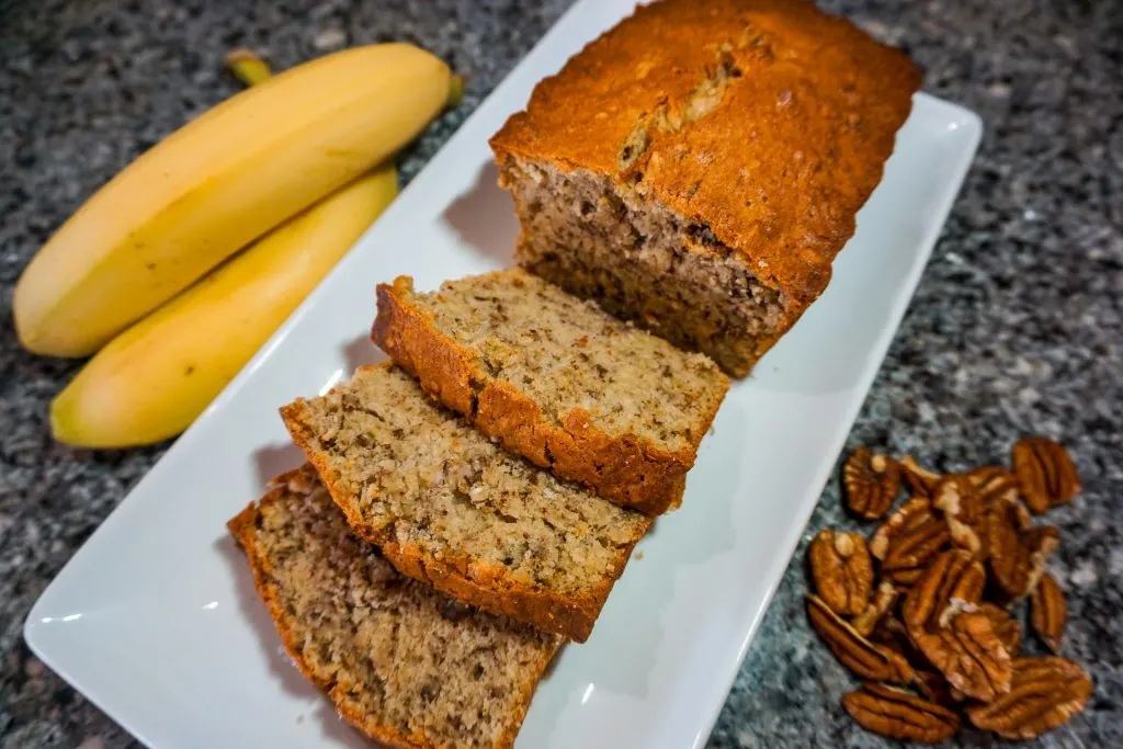 A loaf of the best banana nut bread with three slices on a rectangle plate. On the side are bananas and pecan halves.