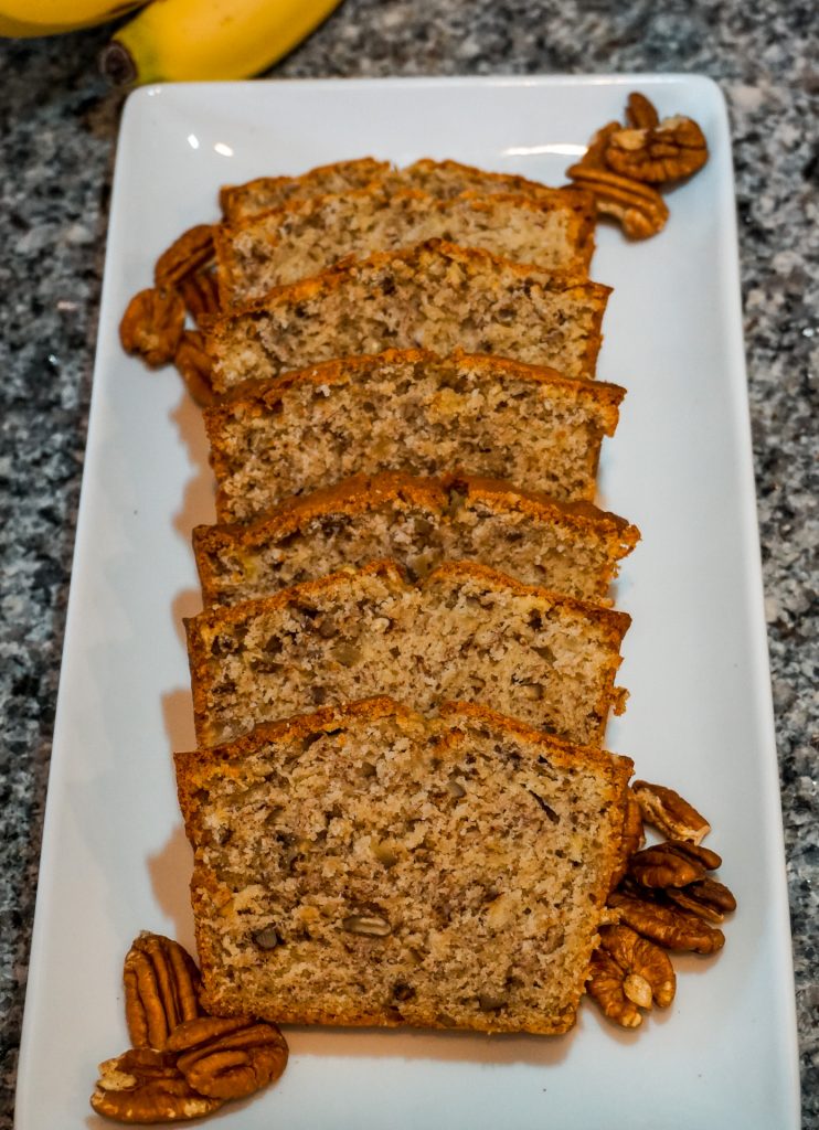 Several slices of banana nut bread in a row on a white rectangle plate with pecan halves.