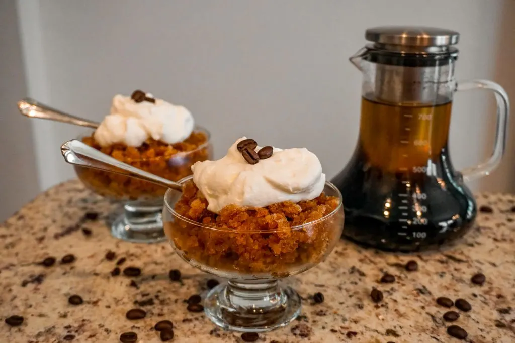 A clear glass coffee maker filled with cold brew coffee next to two bowls filled with cold brew coffee granita and a dollop of whipped cream on top.