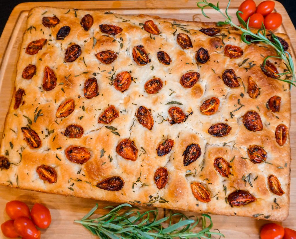 A beautiful loaf of roasted cherry tomato and rosemary focaccia bread on top of a wooden board with fresh rosemary springs and cherry tomatoes.