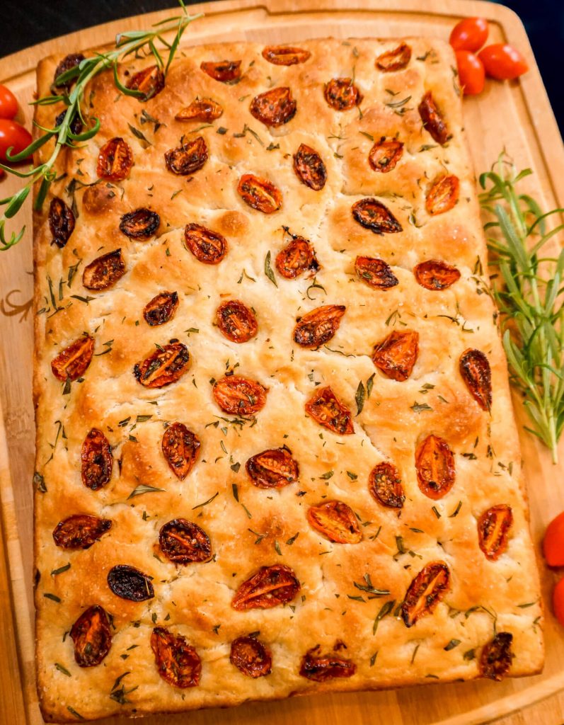 A loaf of roasted cherry tomato and rosemary focaccia bread on a wooden board with fresh rosemary springs and cherry tomatoes surrounding it.
