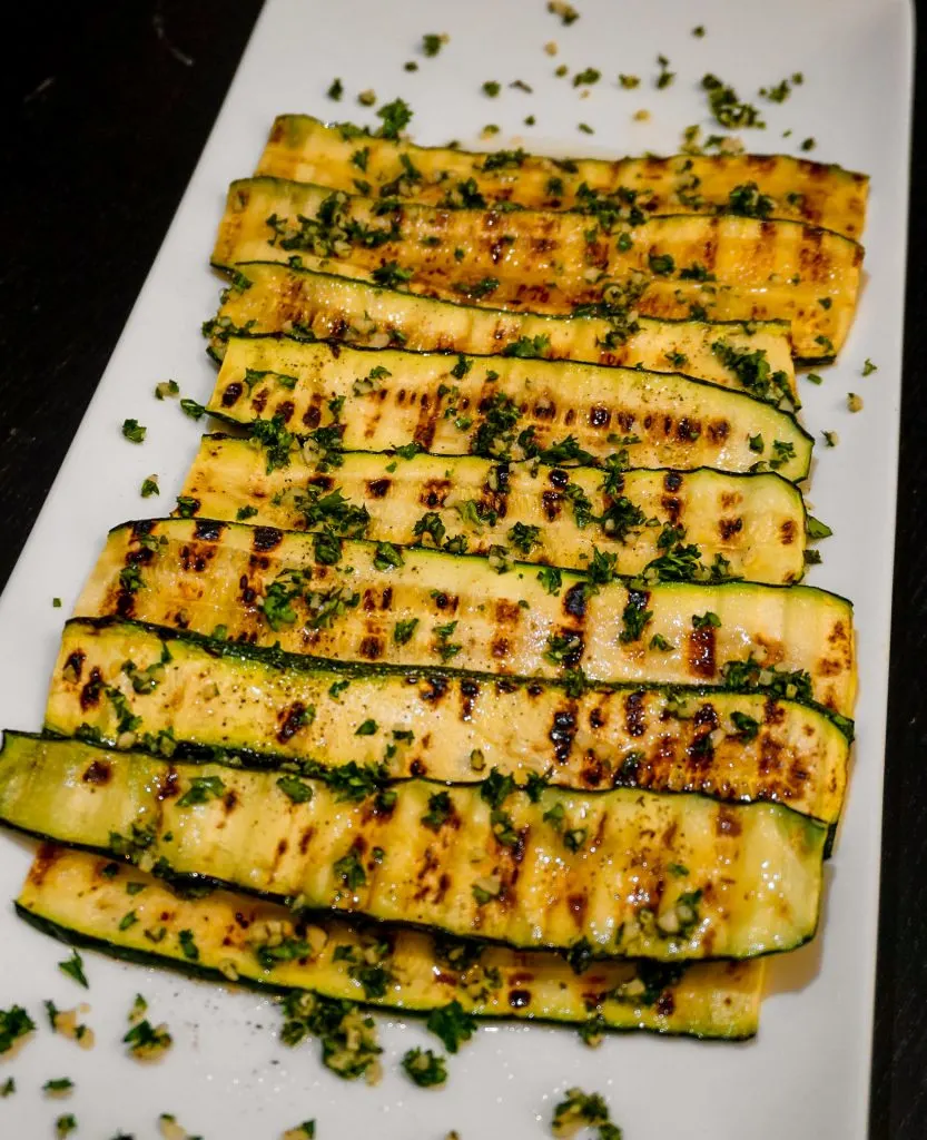 Layers of grilled zucchini with fresh herbs, garlic, and olive oil on a white plate.