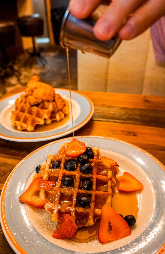 A man pouring maple syrup over Belgian waffles with fresh blueberries and strawberries from Layered in McKinney, Texas.