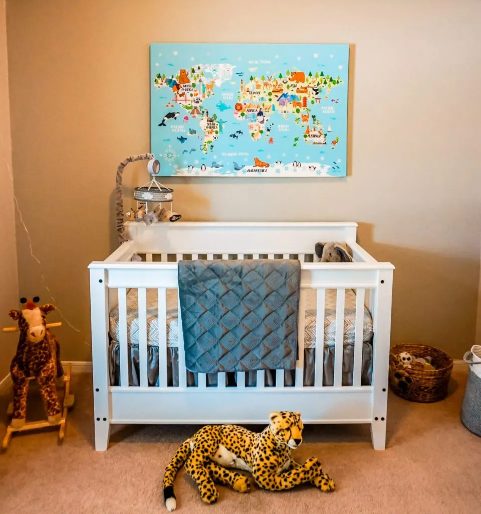 A safari themed nursery with a world map canvas, giraffe rocking chair, cheetah plush toy, white crib with a grey blanket, and more.