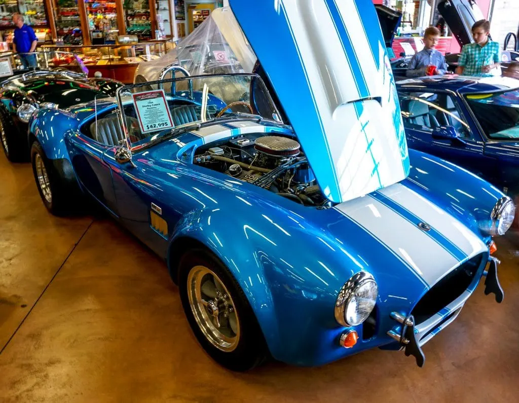 A classic bright blue Shelby Cobra convertible with white stripes in Fast Lane Classic Car's garage.