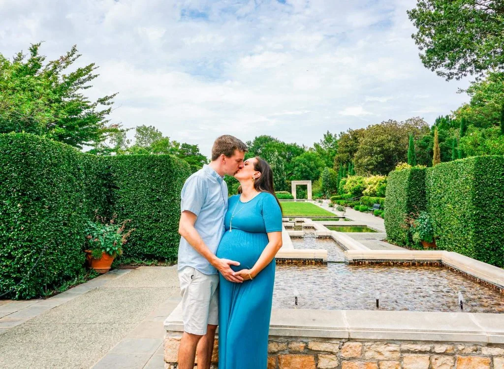 An expecting couple kissing in a beautiful garden at the Dallas Arboretum for their maternity photoshoot. 