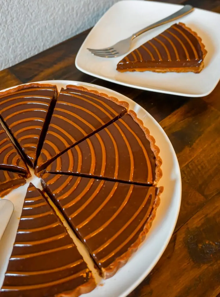 Half of a chocolate caramel tart sliced individually with a plate of one slice in the back.