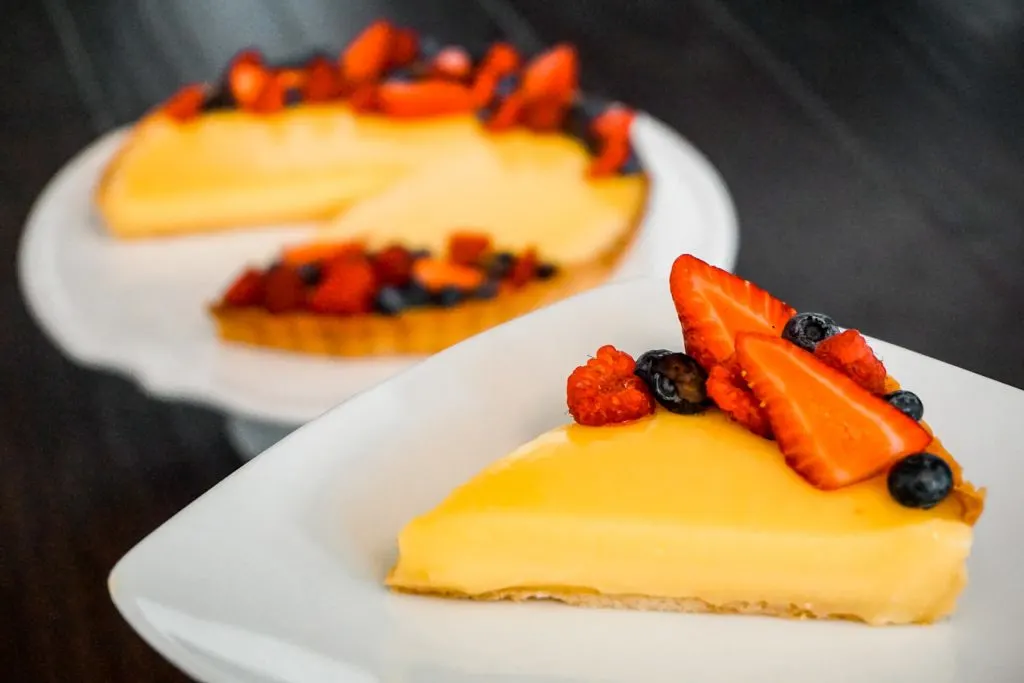 A slice of French Lemon Tart (Tarte au Citron) with fresh berries in focus with the whole tart in the background.