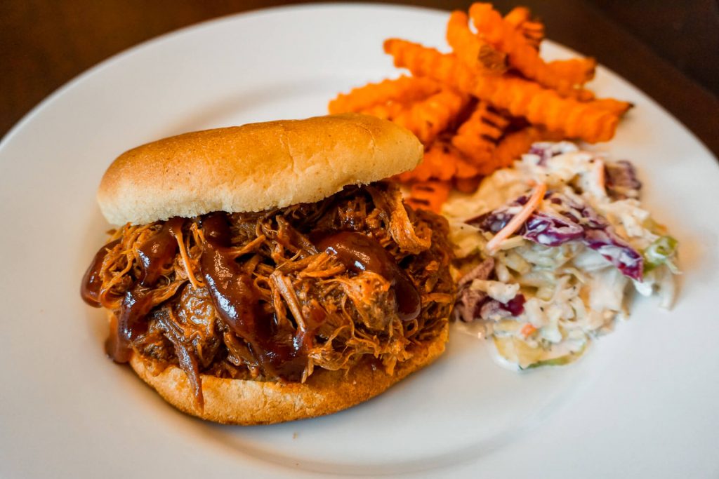 A plate of Texas pulled pork sandwich with a side of coleslaw and sweet potato fries. 