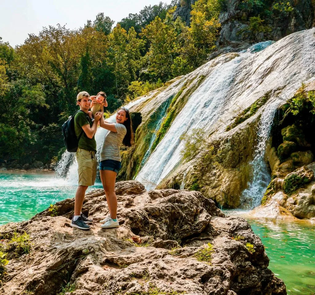 A couple holding up their baby boy on a rock in front of Turner Falls' 77 ft. waterfall.