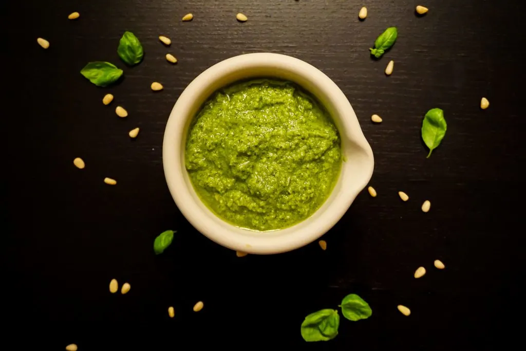 A bowl of bright green pesto with pine nuts and small basil leaves surrounding it.