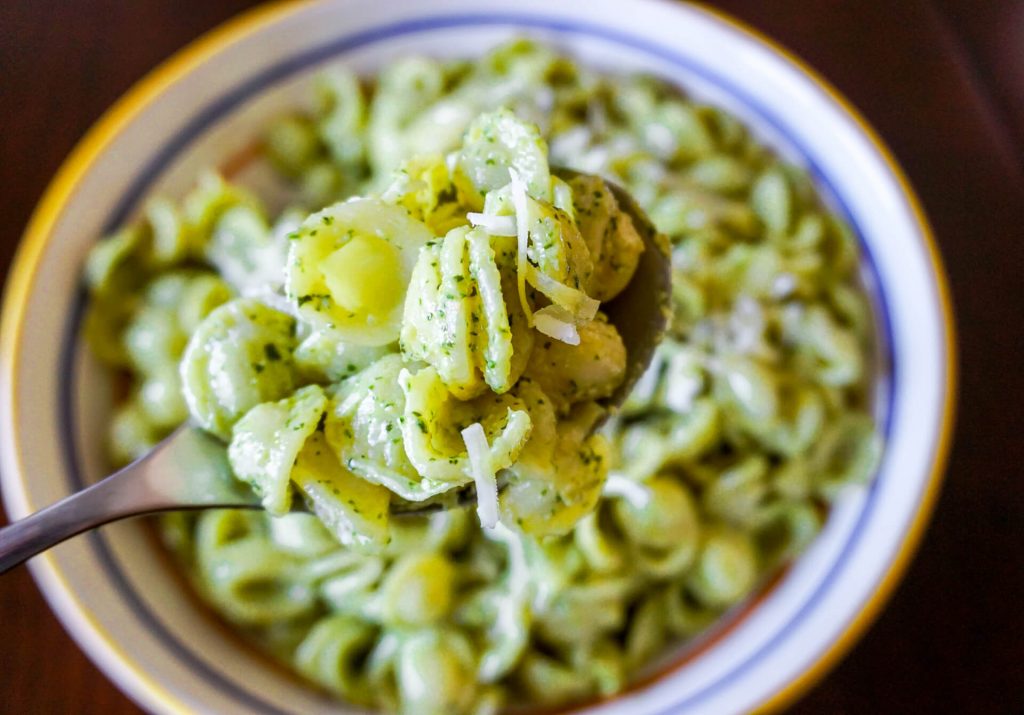A spoon of pesto orecchiette close up with a big bowl of it blurred behind the spoon.