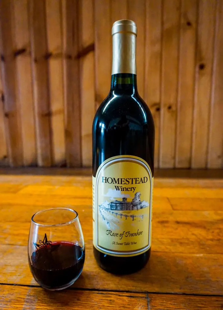 A bottle of Homestead Winery's award-winning wine, Rose of Ivanhoe, alongside a stemless glass of red wine.