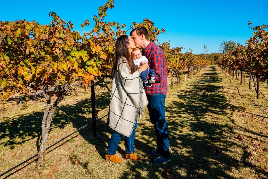 A couple holding their baby between them while kissing in a vineyard in North Texas.