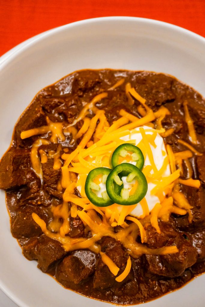 A bowl of Texas Chili with chunks of beef chuck, shredded cheddar cheese, sour cream, and sliced jalapeños. 