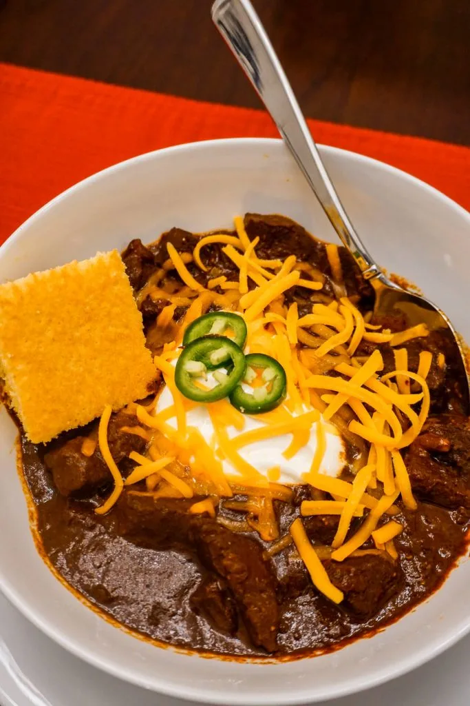 A bowl of authentic Texas Chili with a slice of cornbread, dollop of sour cream, sliced jalapeños, and shredded cheddar cheese. 