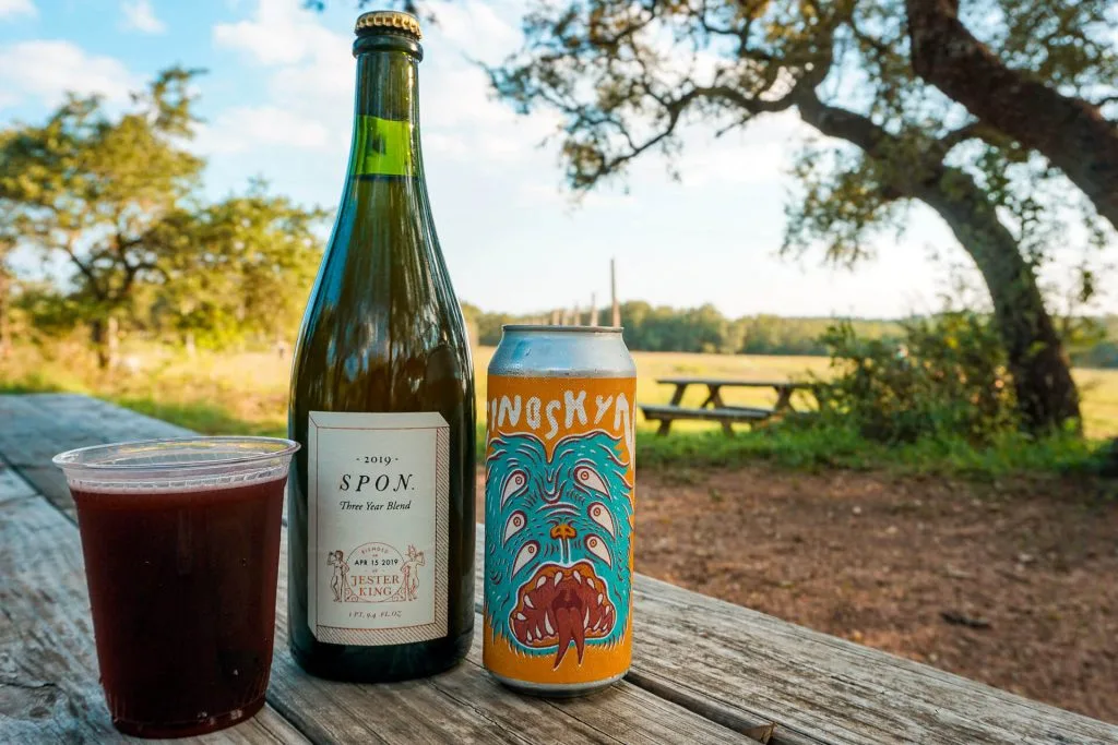 A plastic cup of beer, bottle of Spon beer, and can of beer on a wooden picnic table at Jester King Brewery.